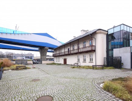 Kolorowa fotografia przedstawiająca budynek Muzeum Podgórza oraz jego najbliższe otoczenie. W tle wiadukt.