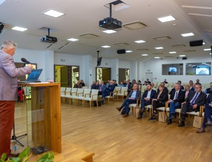 Kolorowa fotografia. Mężczyzna przemawiający na mównicy. Grupa słuchających osób.